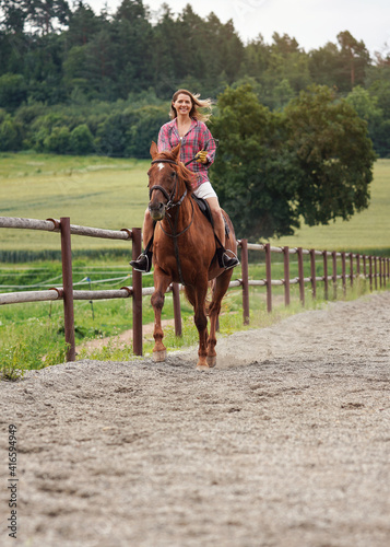 Young woman wearing shirt riding brown horse in sand paddock by wooden fence, hair moving in air because of speed