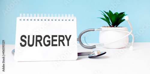 The text SURGERY is written on notepad near a stethoscope on a blue background. Medical concept
