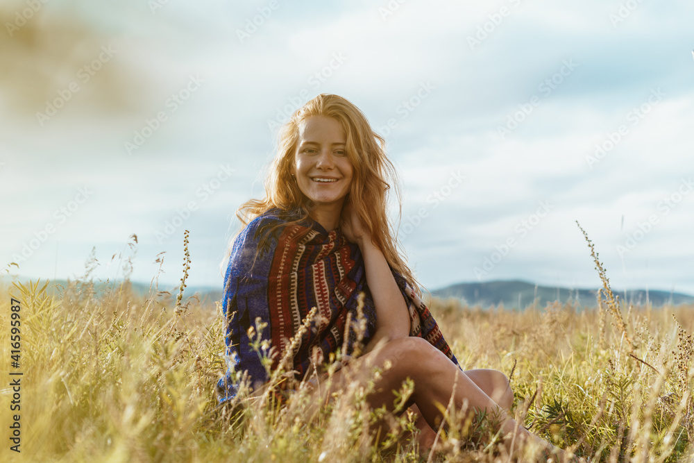Obraz premium among the grass in the meadow sits a red-haired girl with bare legs wrapping her shoulders with a colored cape. High quality photo