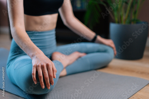 Close-up view of unrecognizable young woman meditating at home sitting in lotus position on yoga mat holding hands on knee. Concept of sports training.