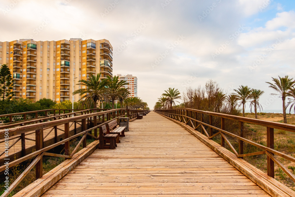 Wooden promenade on Gandia Beach, with wooden benches, palm trees on the sand of the beach, and some tourist apartments, on an afternoon with cloudy skies.