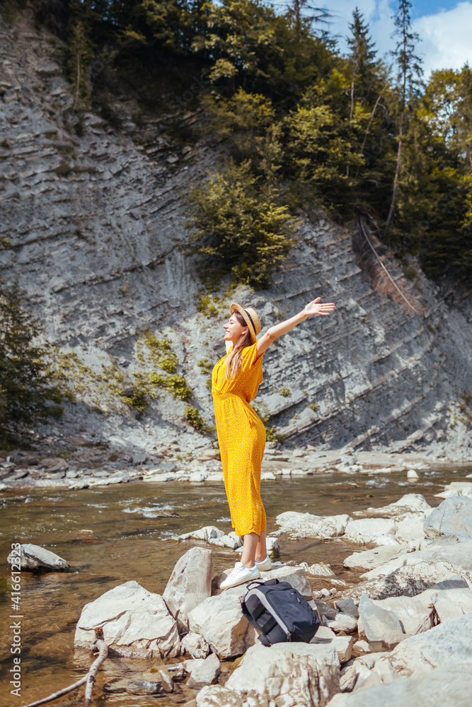 Obraz premium Woman tourist walking by mountain river enjoying landscape. Traveler feels happy raising arms. Summer vacation