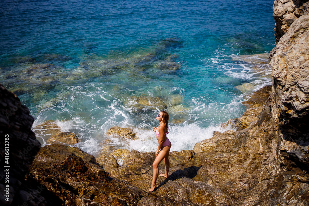 Young beautiful woman in a swimsuit stands on a rocky beach of the Mediterranean Sea. The concept of sea recreation. Selective focus