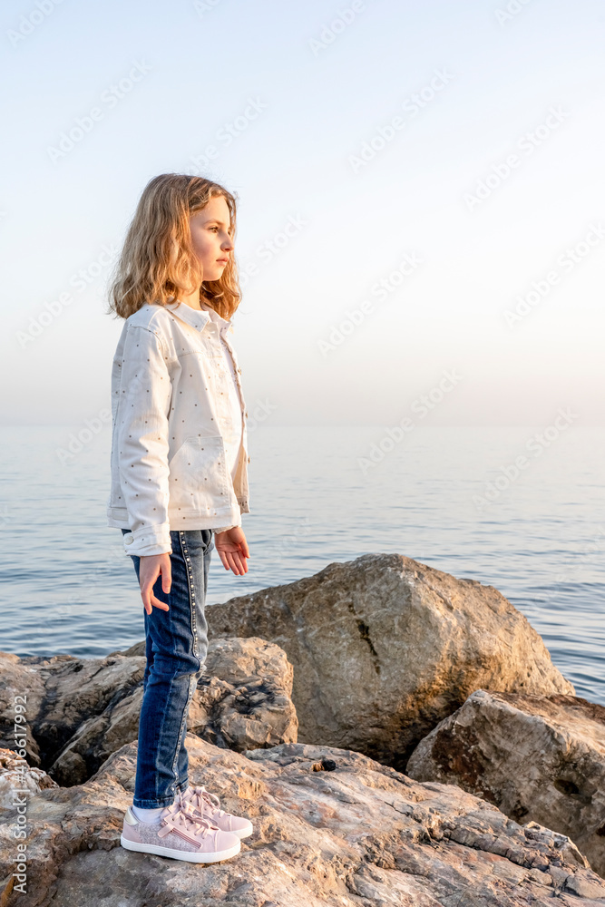 Beautiful girl stands on the stones by the sea and looks away. Warm evening sunset in pastel color. High quality photo