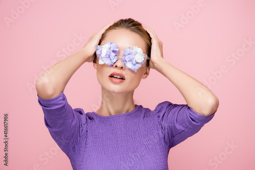 young woman with flowers in eyeglasses touching head while posing isolated on pink © LIGHTFIELD STUDIOS