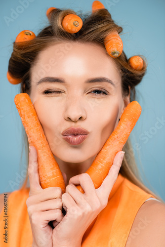 cheerful woman with carrots instead of curlers winking and sending air kiss isolated on blue