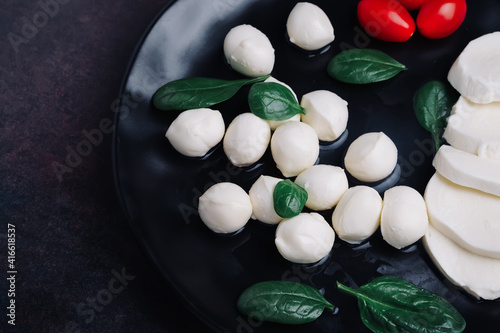 White small mozzarella cheese balls  spinach leaves and tomatoes on black plate.