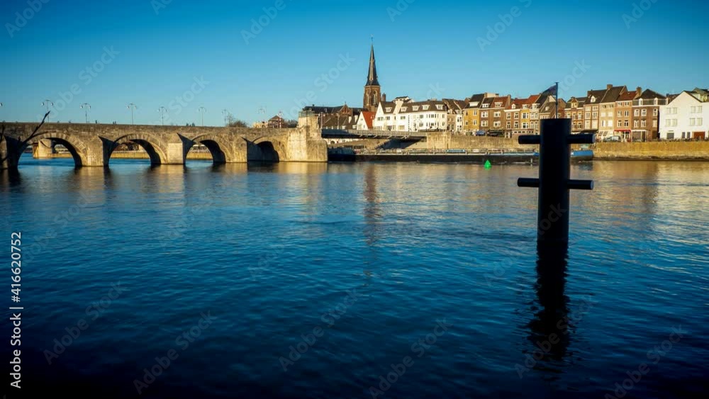 Time lapse day view of a freight ships passing the old town of Maastricht, the Netherlands, with people walking along the promenade in front of old town houses reflecting at the river Maas