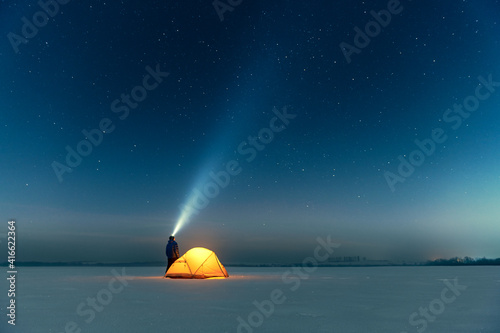 Tourist with flashlight near yellow tent lighted from the inside against the backdrop of incredible starry sky. Amazing night landscape. Tourists camp in snowy field. Travel concept photo