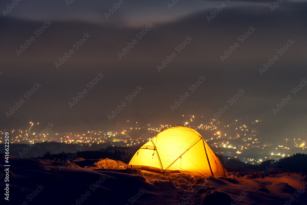 Yellow tent lighted from the inside against the backdrop of glowing city lights in fog. Amazing snowy landscape. Tourists camp in winter mountains. Travel concept