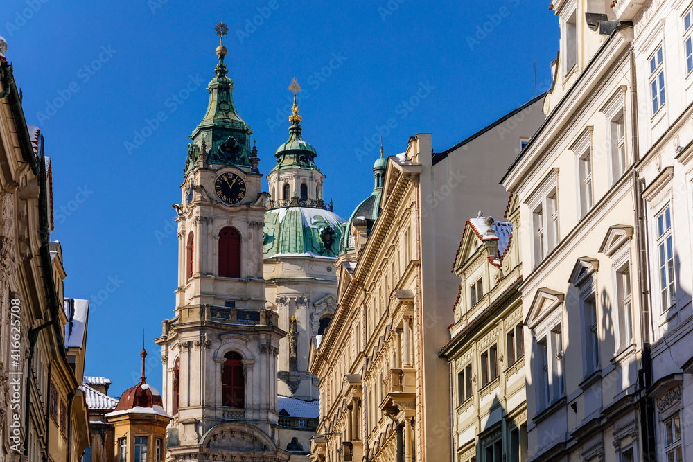 View of Baroque Church of Saint Nicholas, green dome and bell tower with clock, sunny winter day, snow on red roofs, Mala Strana or Lesser Town district, Prague, Czech Republic