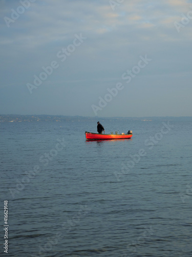 A tiny orange boat sails alone on the calm lake. Evening scene with complementary colors. Fisherman comes home.
