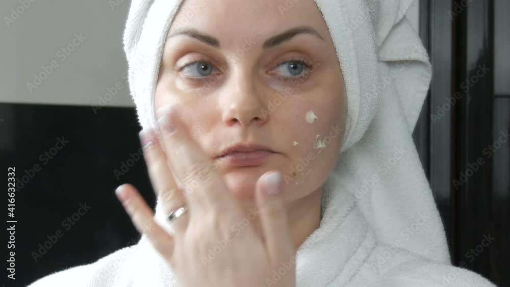 Young beautiful woman applies cream to her face in the bathroom, with a white towel on her head and in a white bathrobe. Facial skin care, prevention of early aging and wrinkles. Young beautiful woman applies cream to her face in the bathroom, with a white towel on her head and in a white bathrobe. Facial skin care, prevention of early aging and wrinkles.
