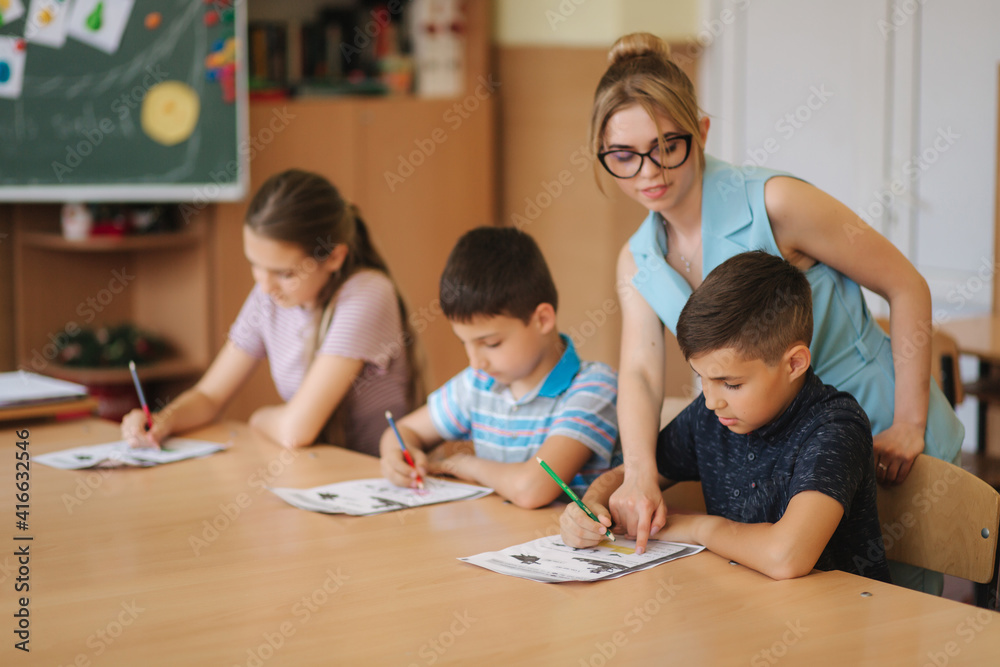 Fototapeta premium Teacher helping school kids writing test in classroom. education, elementary school, learning and people concept. Two boys and one girl stay after school