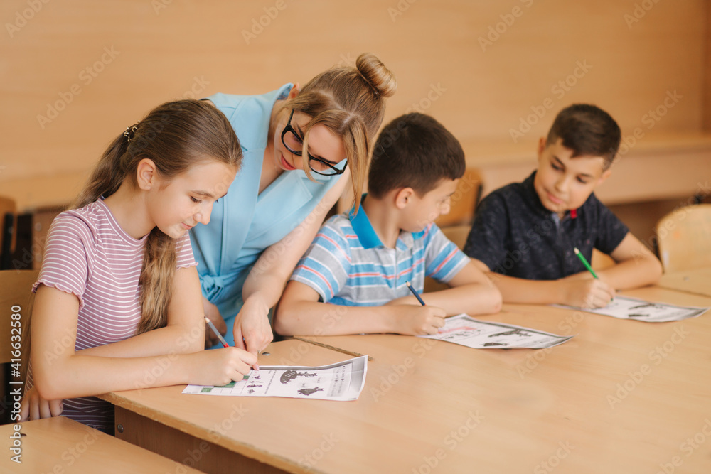 Teacher helping school kids writing test in classroom. education, elementary school, learning and people concept. Two boys and one girl stay after school Teacher helping school kids writing test in classroom. education, elementary school, learning and people concept. Two boys and one girl stay after school