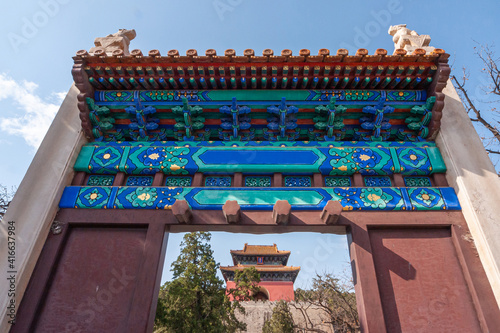 Beijing, China - April 28, 2010: Ming Tombs Changling. Closeup of brown gate with colorful sculpted and painted upper part under blue cloudscape. Red Chinese hall building in back. photo