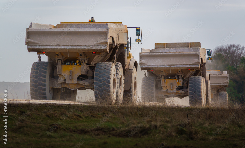 Obraz premium Three yellow Volvo A40E and A40F articulated dump truck earth movers in a convoy kicking up dust as they navigate across Salisbury Plain, Wiltshire UK
