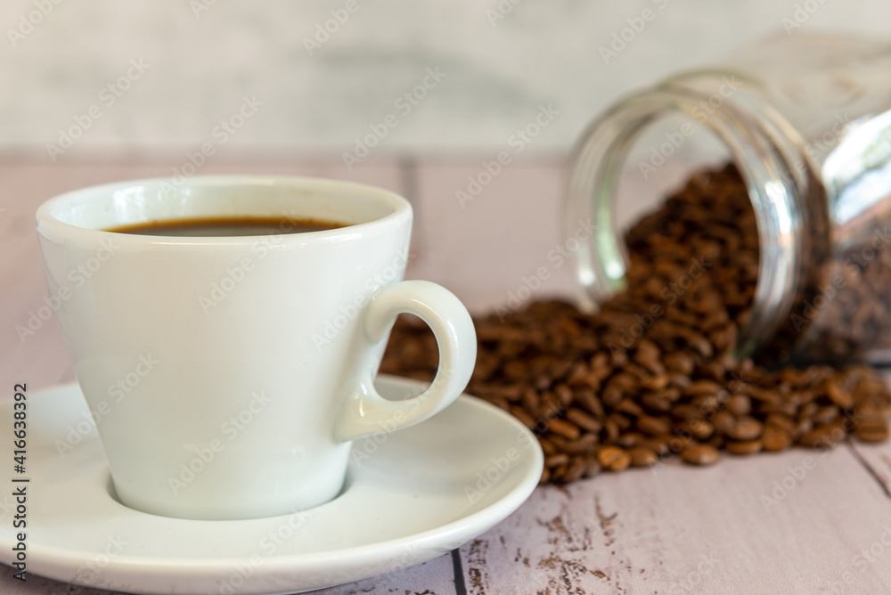 Frasco de vidrio con semillas de cafe tostado desparramadas sobre una mesa, acompañando a una taza de cafe Frasco de vidrio con semillas de cafe tostado desparramadas sobre una mesa, acompañando a una taza de cafe