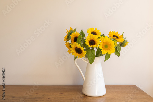 Close up of sunflowers in white jug on oak table against beige wall (selective focus)