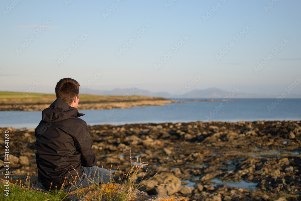 Young man wearing a black raincoat sat on the grass contemplating the coast with a low tide in the evening