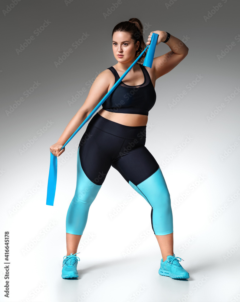 Strong girl working with resistance band. Photo of model with curvy figure in fashionable sportswear on grey background. Sports motivation and healthy lifestyle Strong girl working with resistance band. Photo of model with curvy figure in fashionable sportswear on grey background. Sports motivation and healthy lifestyle