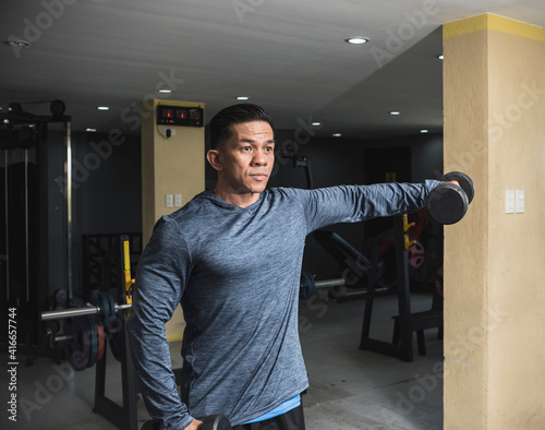 A fit asian man in a sweatshirt does one arm side lateral raises. Shoulder workout and training. Open air gym setting. photo