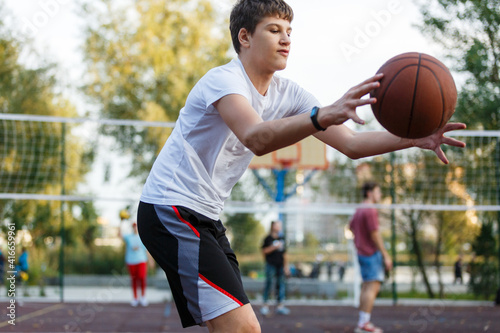 Cute young boy plays basketball on street playground in summer. Teenager in white t-shirt with orange basketball ball outside. Hobby, active lifestyle, sports activity for kids. © Natali