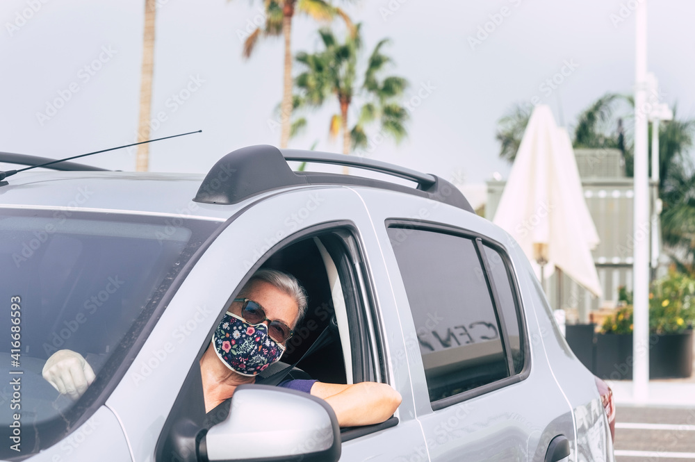 Senior woman wearing a flowered surgical mask due to the covid19 coronavirus ready to drive with his silver looking at camera from the window - active retired people concept