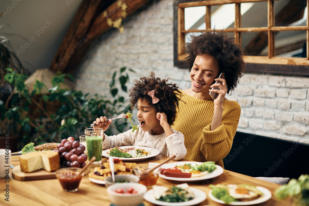 Happy black mother talking on the phone while having breakfast with her daughter in dining room.