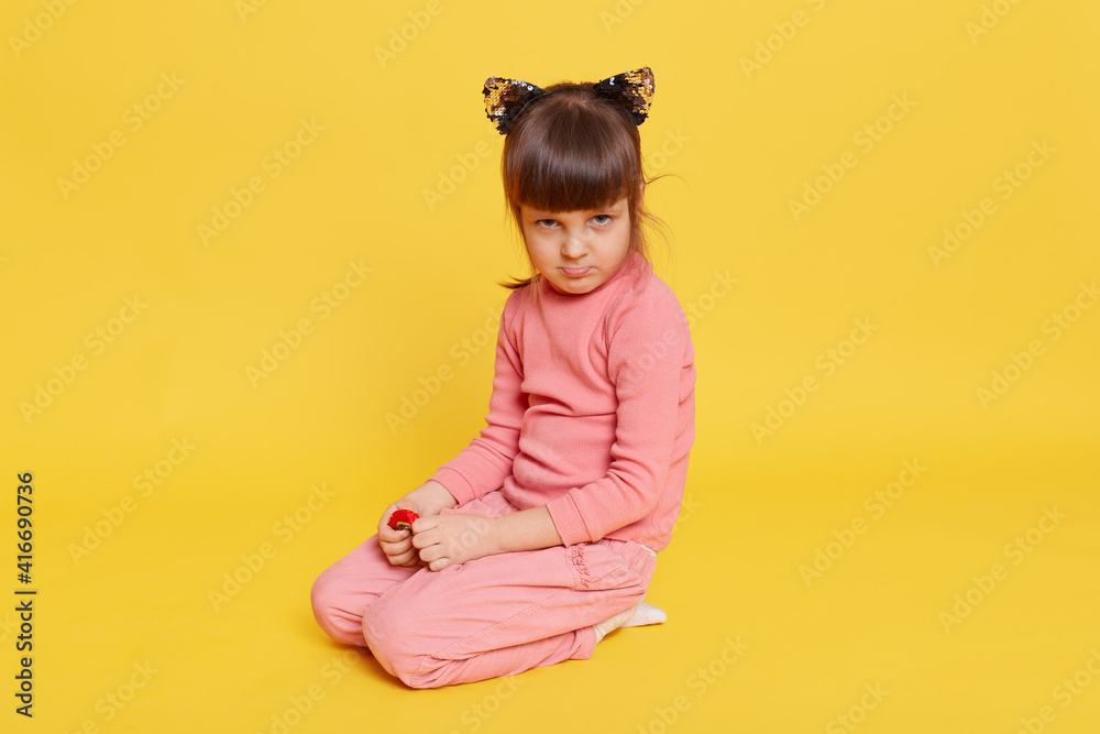 Little dark haired girl looking frowned, sitting on floor against yellow background, toddler wearing casual clothing, offended small kid being upset. Little dark haired girl looking frowned, sitting on floor against yellow background, toddler wearing casual clothing, offended small kid being upset.