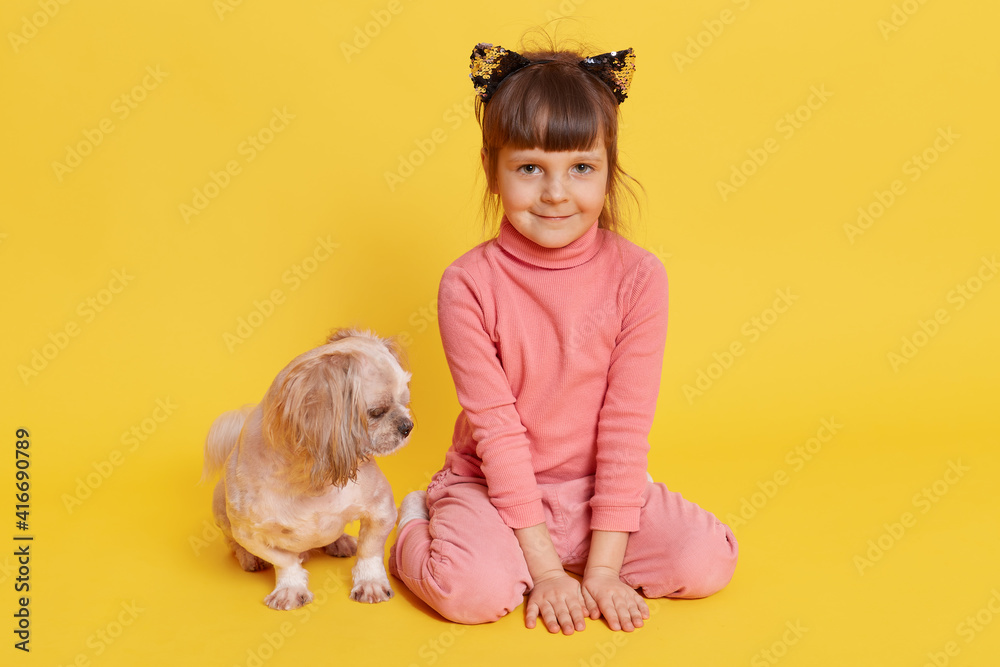 Girl with puppy isolated over yellow background, child looking directly at camera with smile, kid wearing rose sweater, pants and hair band with cat's ears, small kid with Pekingese. Girl with puppy isolated over yellow background, child looking directly at camera with smile, kid wearing rose sweater, pants and hair band with cat's ears, small kid with Pekingese.