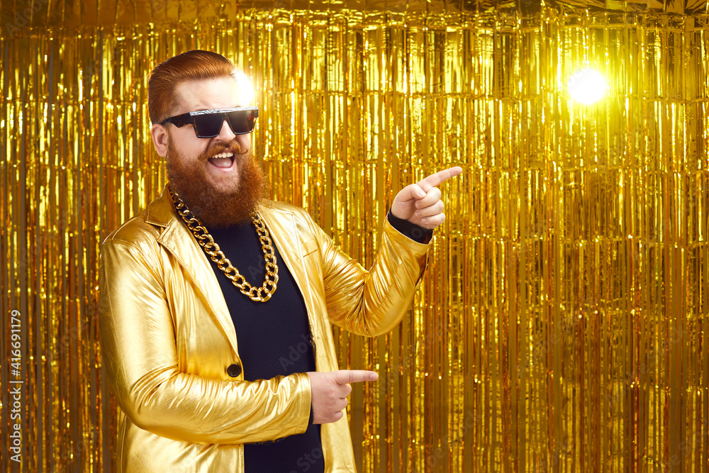 Happy smiling rich man in extravagant golden party jacket, gold chain and  cool glasses with ginger beard and mustache pointing with finger to  advertising copy space. Show, event, concert ad background Stock