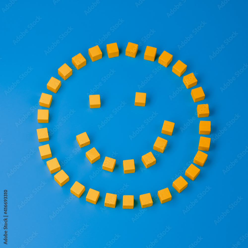 Fototapeta premium Smiley symbol composed of wooden yellow cubes on blue background. Top view, closeup