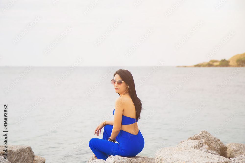 Portrait Asian woman in blue casual wear with sunglasses sitting on the cliff above the sea while traveling in Asia