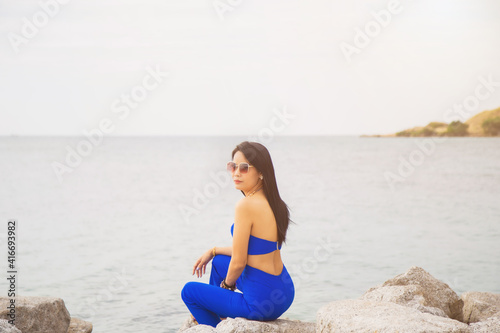 Portrait Asian woman in blue casual wear with sunglasses sitting on the cliff above the sea while traveling in Asia