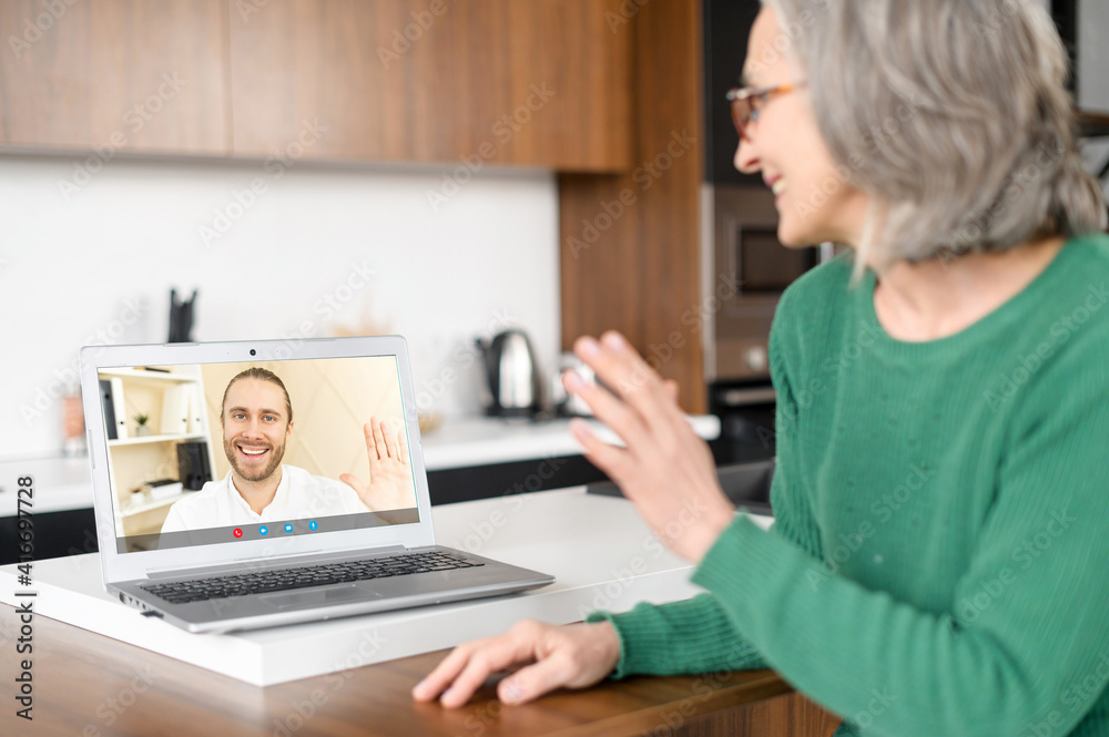 Fototapeta premium Smiling entrepreneur say hello to assistant, aged woman with glasses, sitting at the laptop on the kitchen, from the different subdivisions in the company, can work anywhere, extended capabilities