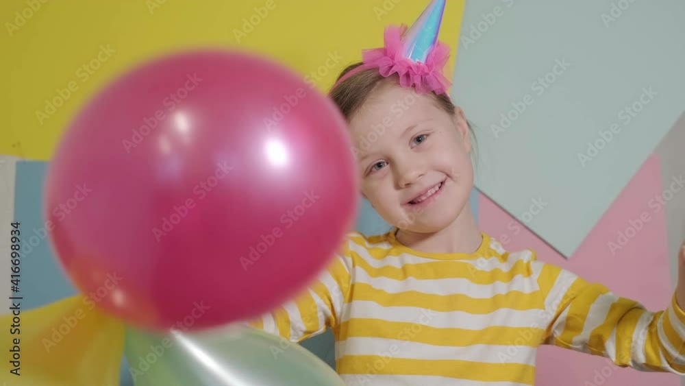 a blonde little girl in a yellow sweatshirt with a pink cap on her head smiles against a colorful background with balloons at a birthday party close up slow motion a blonde little girl in a yellow sweatshirt with a pink cap on her head smiles against a colorful background with balloons at a birthday party close up slow motion