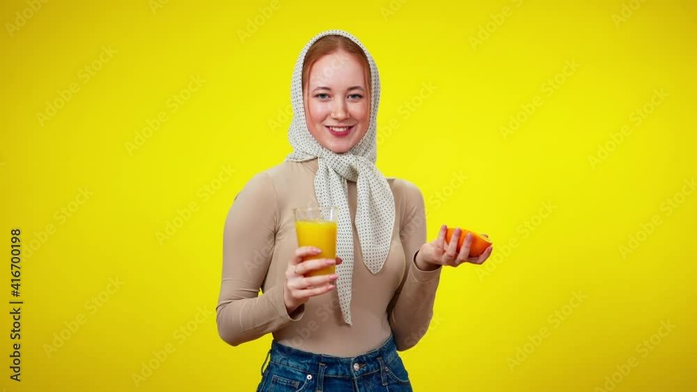Middle shot of charming beautiful young woman holding juice glass and half of orange looking at camera smiling. Portrait of confident vegan Caucasian millennial posing at yellow background. Middle shot of charming beautiful young woman holding juice glass and half of orange looking at camera smiling. Portrait of confident vegan Caucasian millennial posing at yellow background.