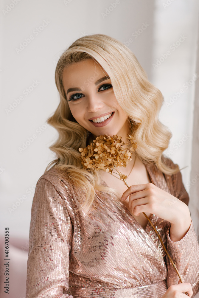 Attractive young blonde woman with curly hair and makeup in a sequined dress in the bright interior of the studio house Attractive young blonde woman with curly hair and makeup in a sequined dress in the bright interior of the studio house