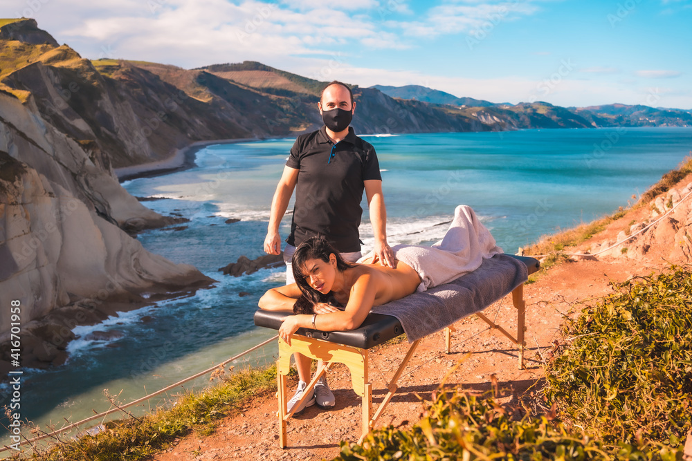 A young man performing a nature massage on the coast near the sea, a dream come true, masseur with face mask in the pandemic