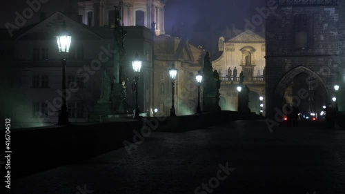 a row of lit lamps on Charles Bridge and a statue of the crucified Jesus Christ on Charles Bridge in Prague from the 17th century and a crossing of the tiles and pedestrian silhouettes
