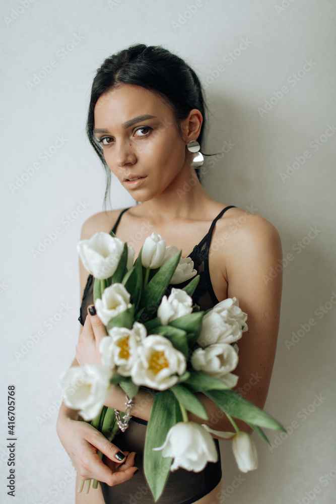 A beautiful brunette girl in a black bodysuit stands sideways and holds a bouquet of white tulips looking at the camera A beautiful brunette girl in a black bodysuit stands sideways and holds a bouquet of white tulips looking at the camera