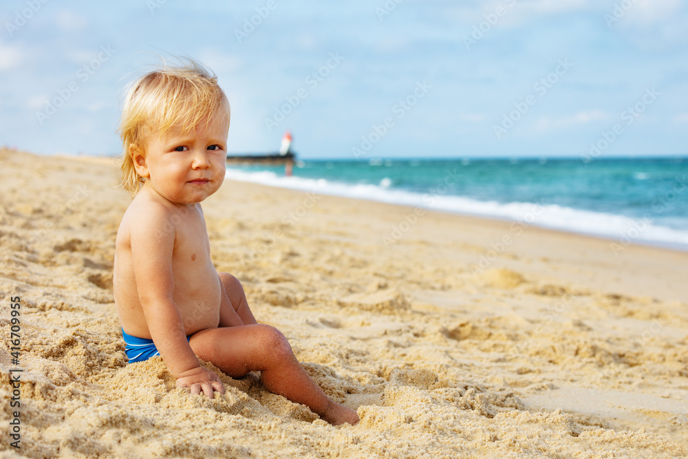 Portrait of a little calm blond toddler boy sitting on the beach near lighthouse during summer sea vacations