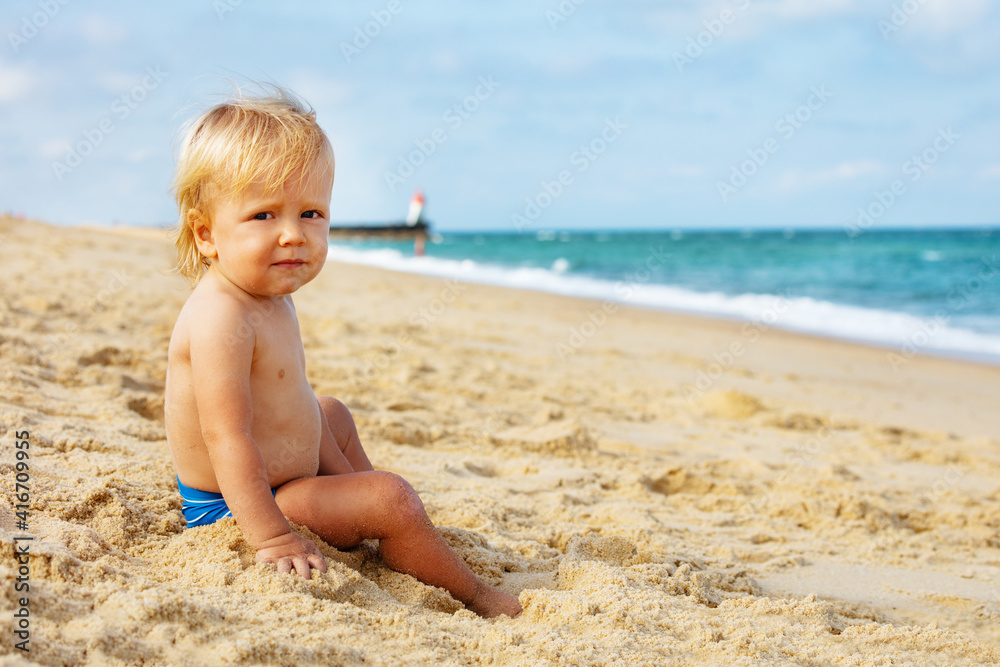 Portrait of a little calm blond toddler boy sitting on the beach near lighthouse during summer sea vacations Portrait of a little calm blond toddler boy sitting on the beach near lighthouse during summer sea vacations
