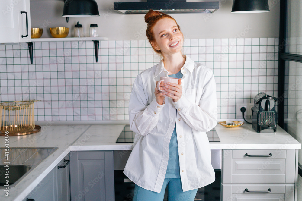 Portrait of dreamy redhead woman holding cup of hot beverage in hands on morning at kitchen. Attractive lady enjoying drinking cup of tea at home. Concept of leisure activity red-haired female. Portrait of dreamy redhead woman holding cup of hot beverage in hands on morning at kitchen. Attractive lady enjoying drinking cup of tea at home. Concept of leisure activity red-haired female.