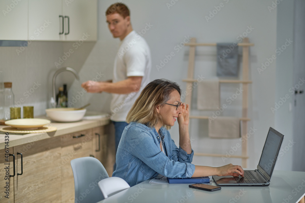 modern multinational couple at home in the kitchen. woman working or studying uses laptop. the husband in the background is cooking or cleaning. healthy relationships without gender stereotypes. modern multinational couple at home in the kitchen. woman working or studying uses laptop. the husband in the background is cooking or cleaning. healthy relationships without gender stereotypes.