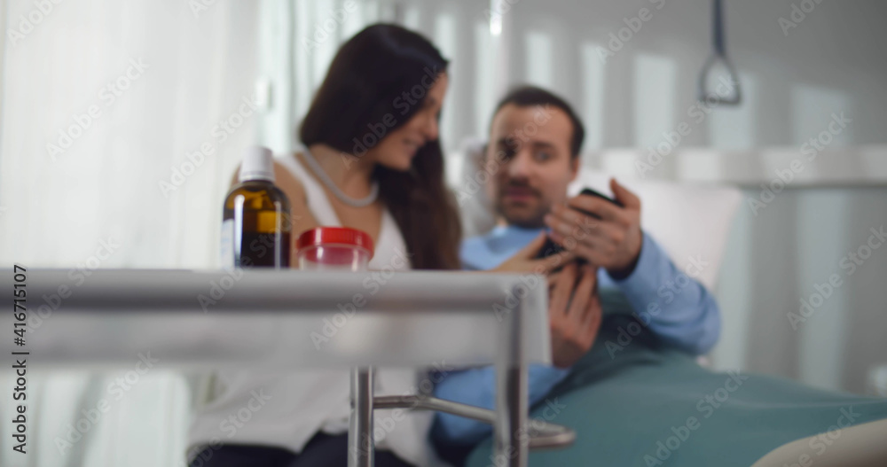 Young woman visiting sick husband in hospital watching photo on smartphone together