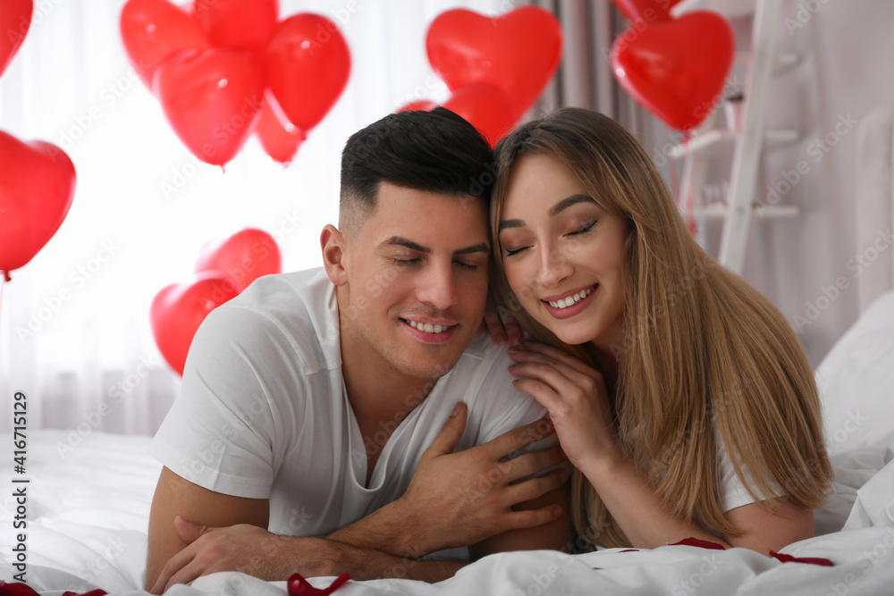 Lovely couple on bed in room decorated with heart shaped balloons. Valentine's day celebration