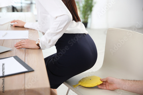 Young man putting whoopee cushion on chair while his colleague sitting down in office, closeup. Funny joke photo