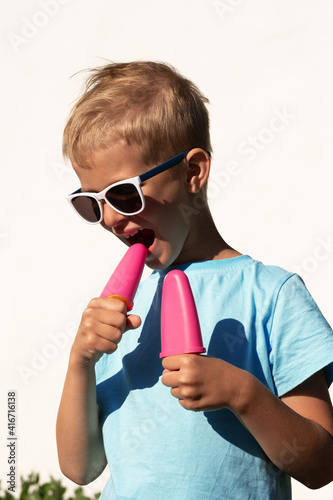 Charming blonde teen in sunglasses bites ice cream on a hot day against the backdrop of a snow-white house